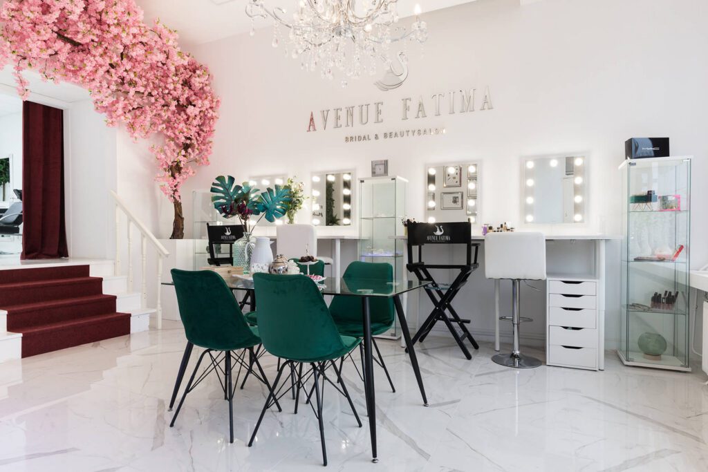 A bright, elegant bridal and beauty salon named "Avenue Fatima". The space features multiple makeup stations with illuminated mirrors, white rolling drawers, emerald green velvet chairs around a glass table, and a decorative pink cherry blossom tree.