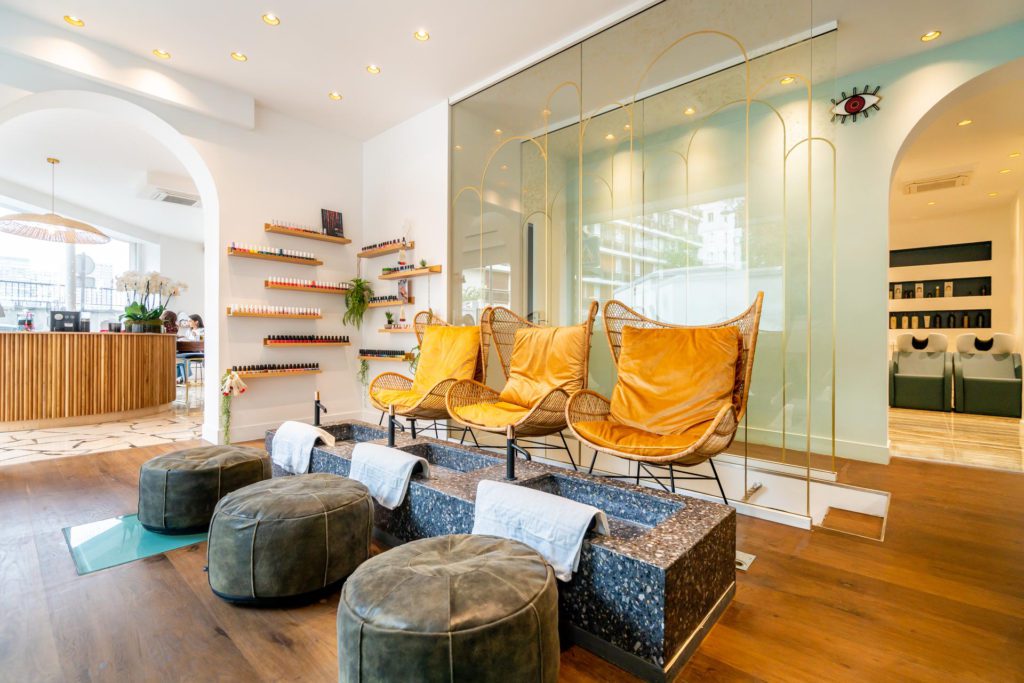 An elegant, brightly lit beauty salon interior featuring a dedicated pedicure station with three mustard-yellow wicker armchairs, stone basin tubs with white towels, and matching grey footstools on a wooden floor.