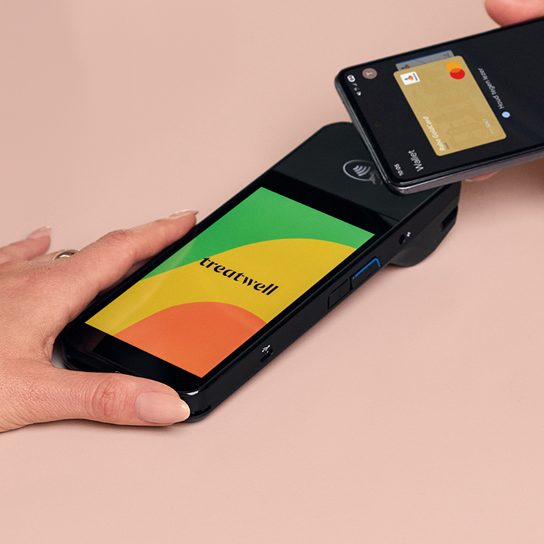 A person holds a black card machine with a multi-coloured screen featuring a green, yellow, and orange design with the dark blue "Treatwell" logo, while another person taps their smartphone against it to make a contactless payment via a digital wallet. The scene is set on a light pink surface.