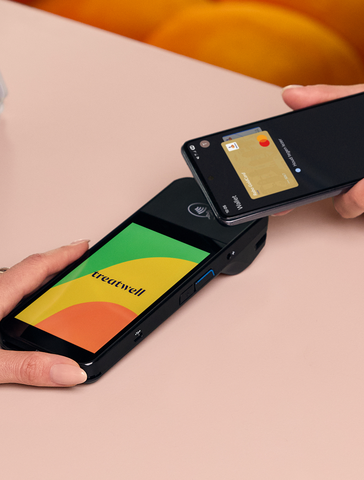 A high-angle close-up of a contactless payment being made. One person holds a Treatwell POS card machine displaying the brand logo, while another person taps a smartphone over it to pay. The background features a bright orange velvet sofa.