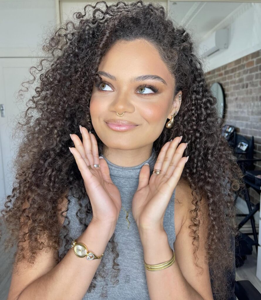 Woman with curly hair and side part in gray sleeveless top, showcasing beauty trend for spring 2026. Credits: @tannia_t_mua