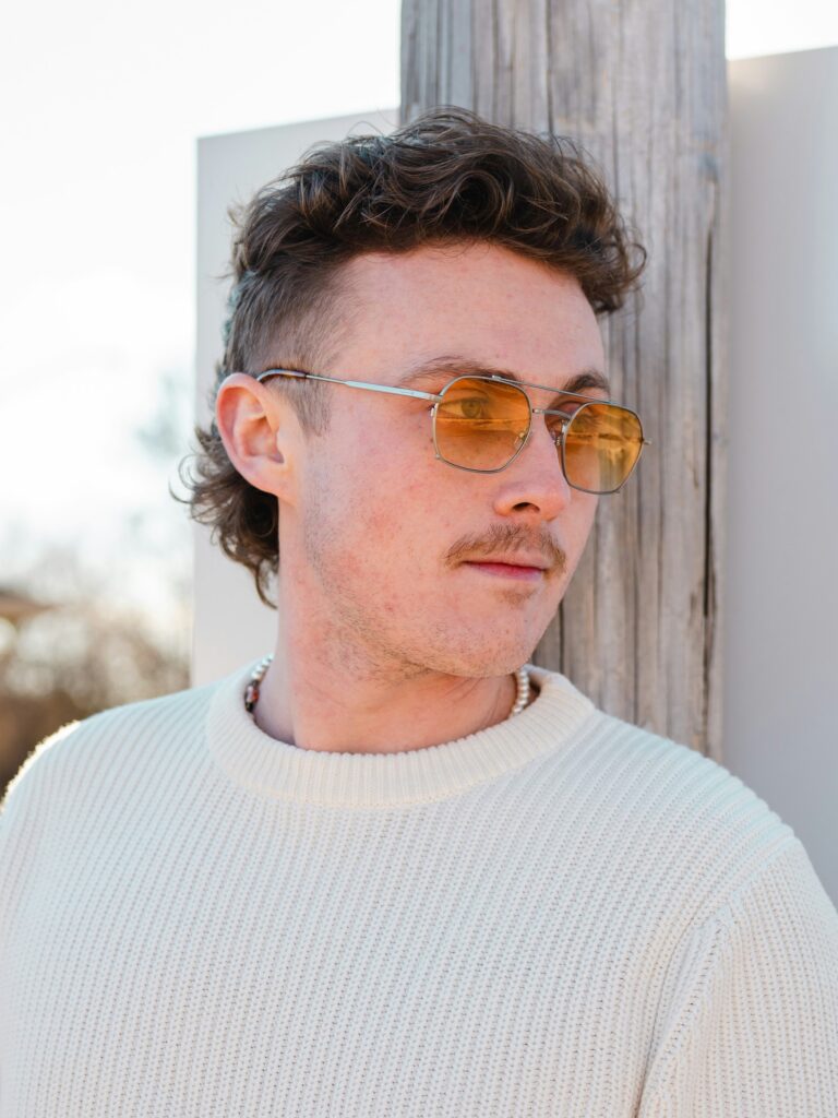 [ALT_TEXT: A young man with a curly mullet haircut leaning on a wall outside on a sunny day.]
