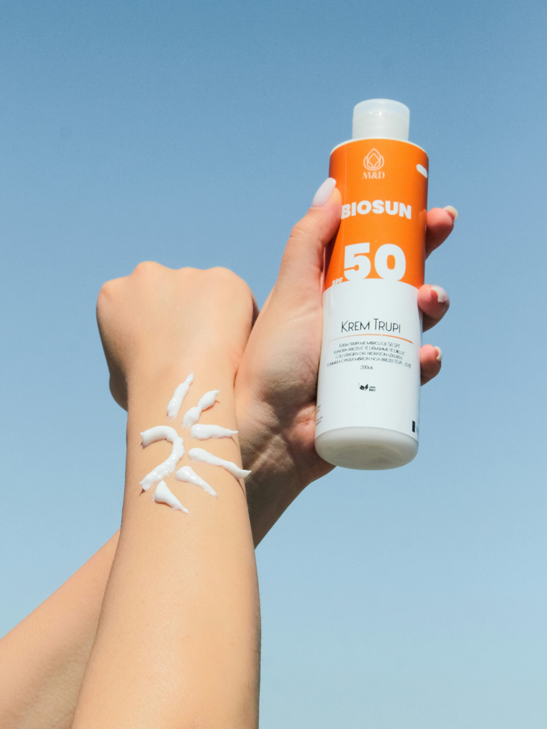 Close-up of a woman's arms against a blue sky, one hand holds a bottle of suncream and one forearm has suncream in the shape of a sun