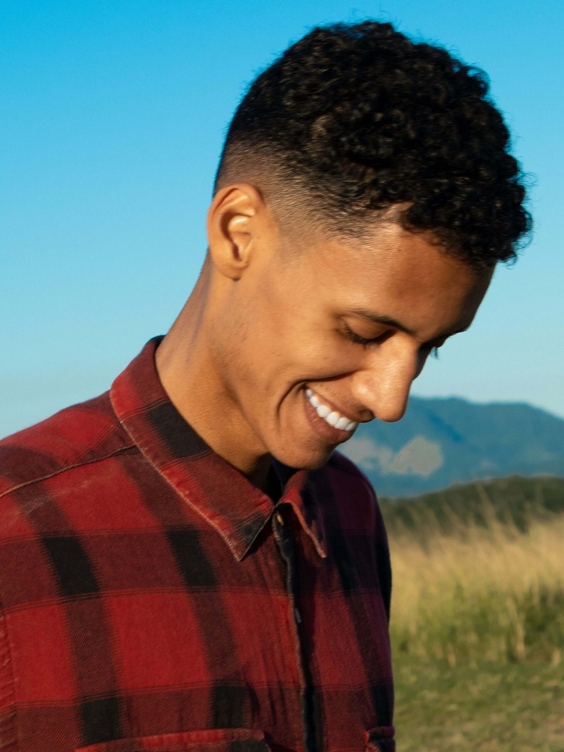 Man smiling outside against a background of mountains and blue sky with curly hair paired with a mid taper fade