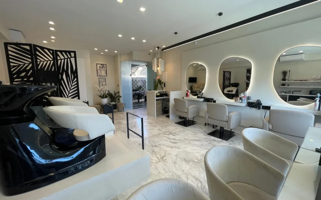 A sleek hair salon with white marble floors, cream leather styling chairs beneath LED-rimmed oval mirrors, a glossy black wash station, and a geometric palm-leaf room divider.