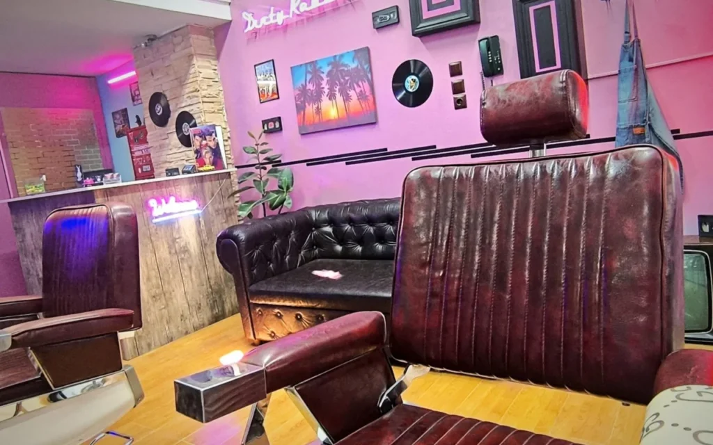 A retro-styled barbershop with pink and purple walls, neon signage, vinyl records mounted as decor, a palm-tree sunset painting, dark leather barber chairs, and a Chesterfield sofa on warm wood flooring.
