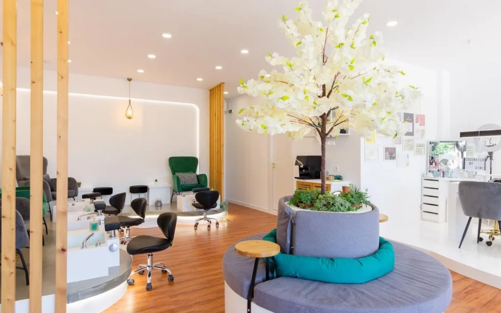 An open, nature-inspired nail salon with a large decorative white blossom tree as the centrepiece, emerald-green velvet armchairs at pedicure stations, wooden slat dividers, and warm wood flooring.