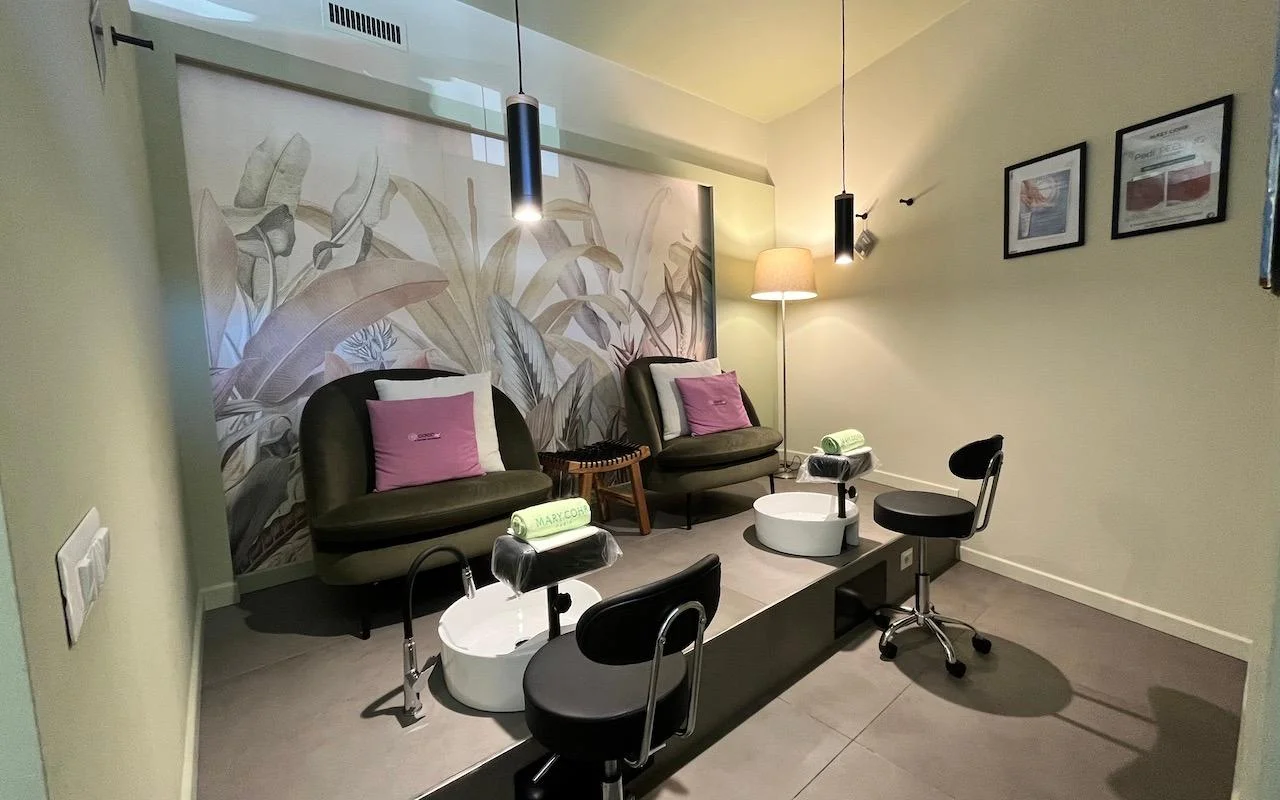 A sophisticated pedicure station with two deep green velvet armchairs and pink cushions. White foot baths are built into a grey raised platform, set against a mural of large tropical leaves on the back wall.