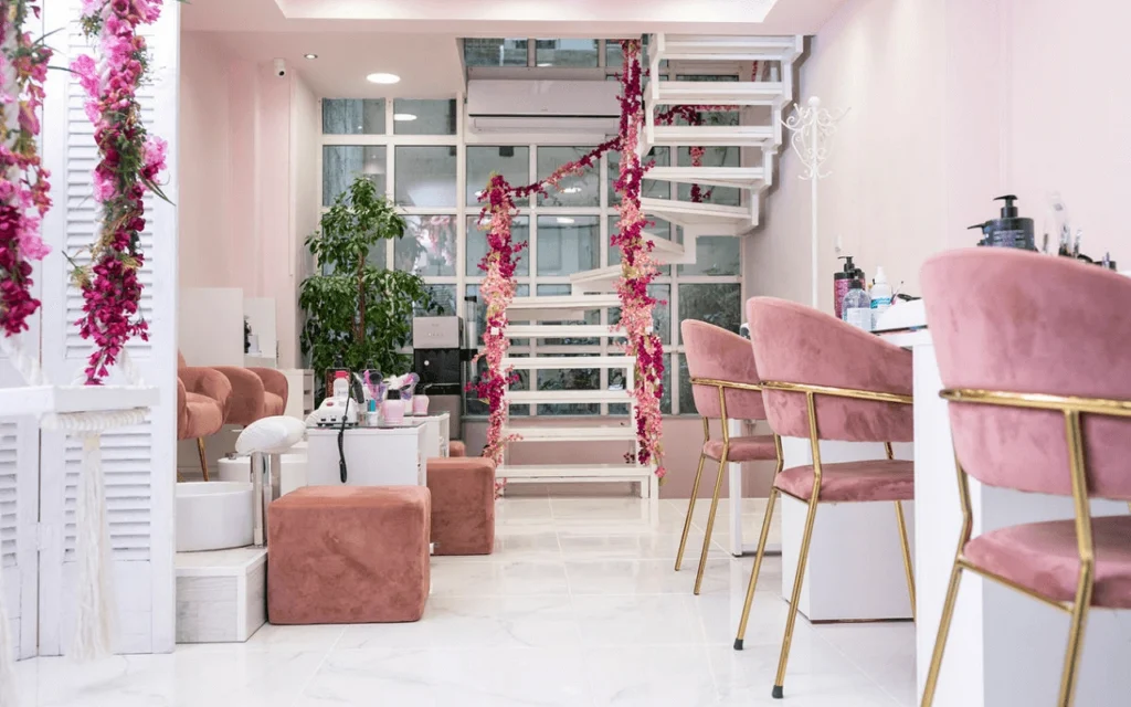 A feminine nail salon with blush-pink velvet chairs, a white spiral staircase draped in pink faux flowers, shuttered dividers, and gold-legged manicure stations on marble-effect flooring.