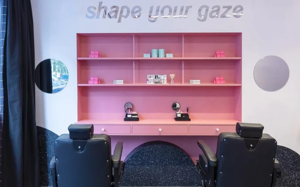 A specialist brow and lash studio with a bold pink shelving unit displaying beauty products, silver "shape your gaze" lettering on the wall, and black leather treatment chairs on a dark speckled floor.
