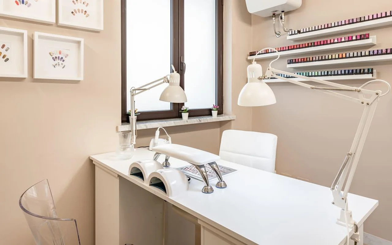 A dedicated nail station at AG The Beauty Room’s with a clean white desk, dual UV lamps, adjustable task lights, and shelves displaying rows of coloured nail polishes. Framed nail art designs hang on the warm-toned walls.