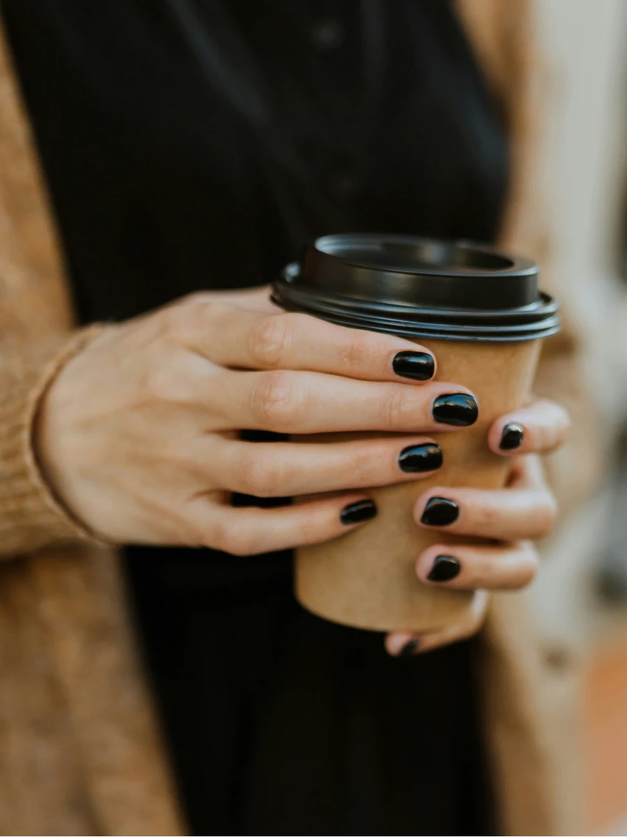 Close-up of a woman with a glossy black gel manicure holding a takeaway coffee cup