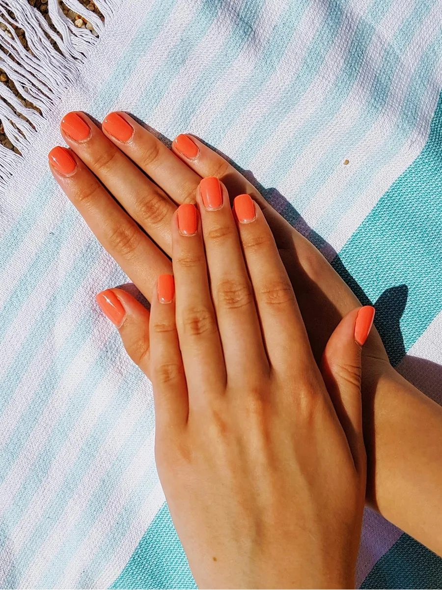Top view of two hands with a shiny orange gel manicure resting on a blue-and-white stripy beach towel