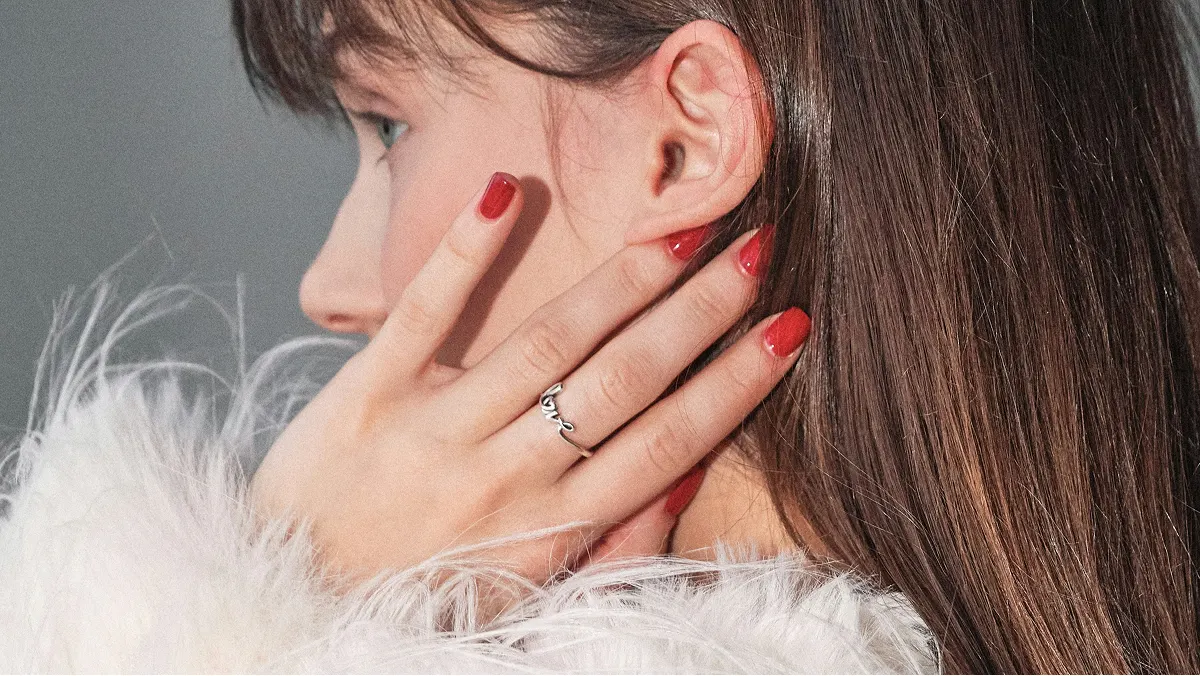 Close-up side view of a brunette woman’s head, her hand touching her face showing a glossy red gel manicure
