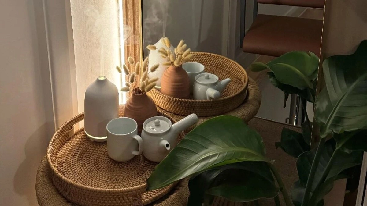 Close-up of a wicker tray holding a diffuser and a tea set next to a large green plant in a salon