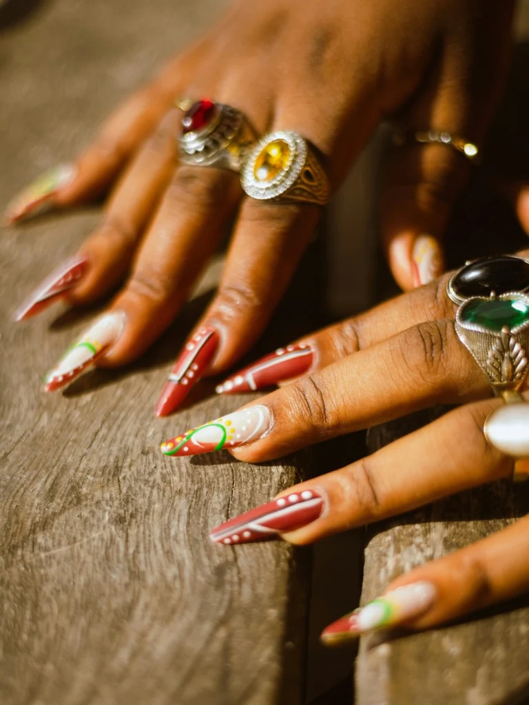 Close-up of a woman’s hands with stiletto-shaped acrylic nails in red and white decorated with green and yellow dots and swirls