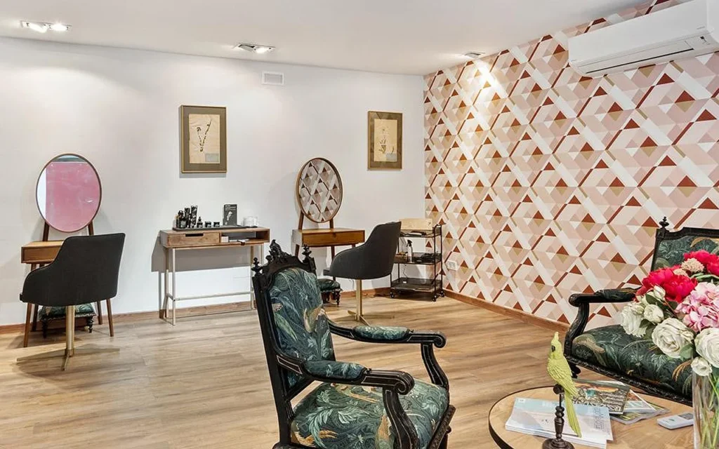 An elegant salon room with a geometric pink and terracotta feature wall, a pair of ornate carved chairs upholstered in green botanical fabric, wooden styling desks with round mirrors, and a vase of pink and red flowers in the foreground.