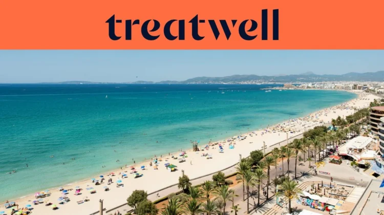 An aerial view of Playa de Palma in Mallorca, with a long curve of golden sand, rows of beach umbrellas, calm turquoise water, and a palm-lined promenade running alongside the city, with hills in the distance under a clear blue sky.
