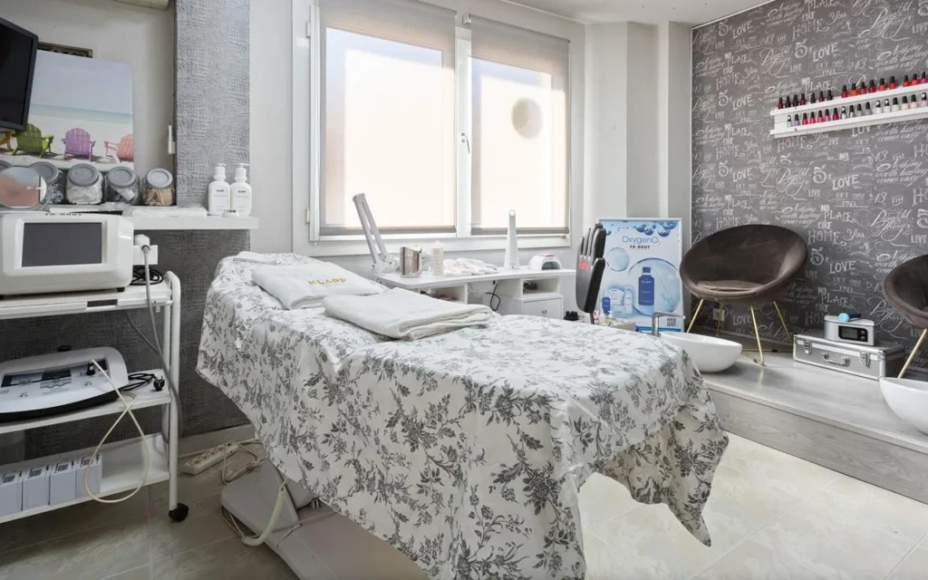 A bright beauty treatment room with a bed dressed in grey floral linen and folded white towels, aesthetics machines on trolleys to the left, a window with the blind half-drawn, and a raised area with a brown velvet chair and pedicure bowls against a script-print feature wall.