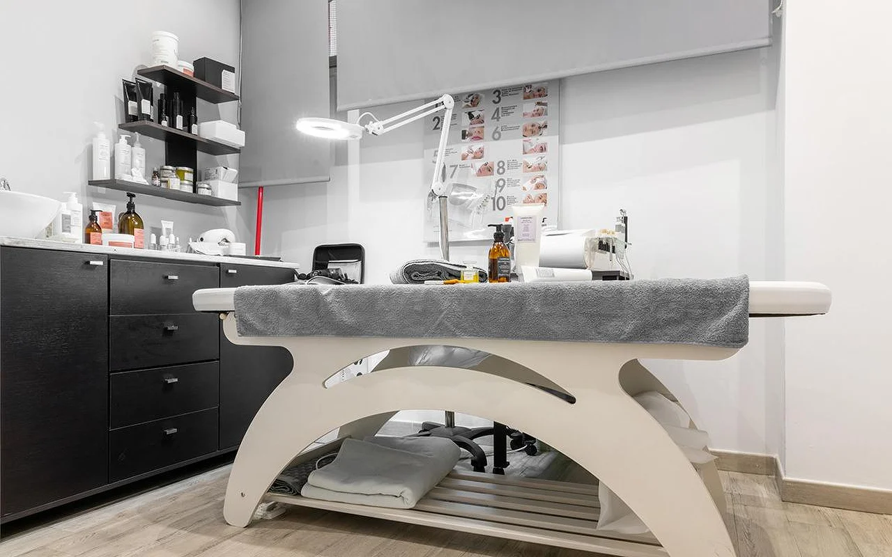 A compact aesthetics treatment room at Atelier Beauty and the Lab, with a cream treatment bed, an adjustable magnifying lamp, dark wall shelves lined with skincare products and a numbered treatment-step chart on the wall.
