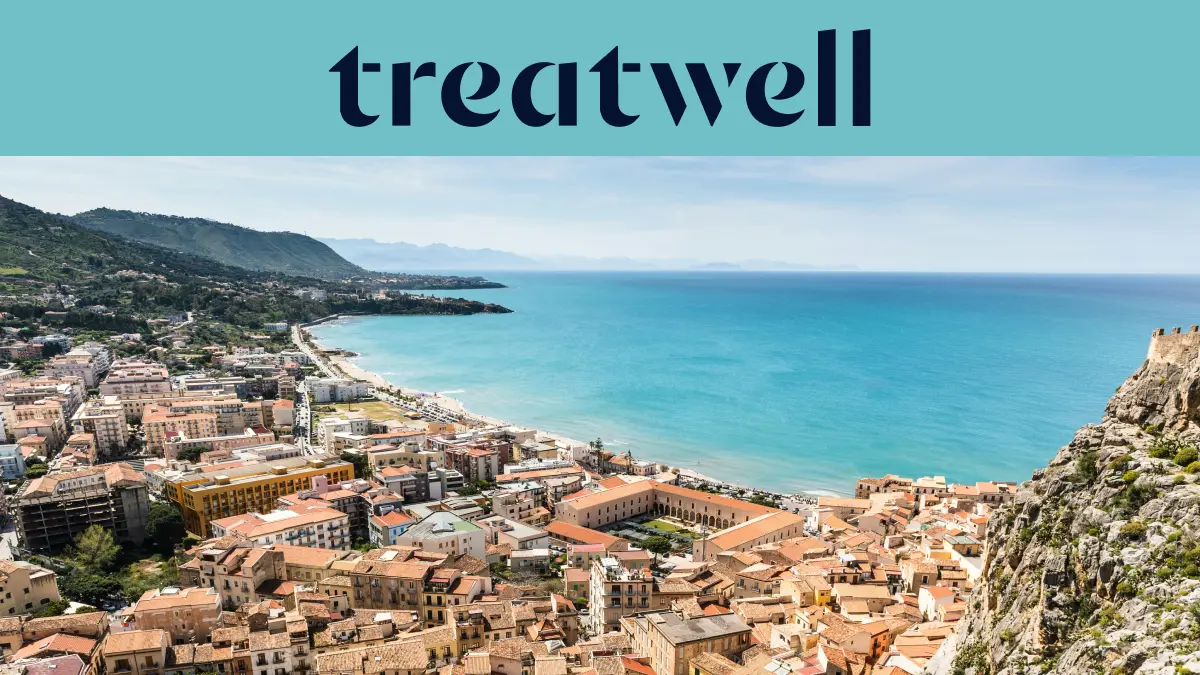 A sweeping view over a Sicilian coastal town, with terracotta-roofed old buildings, a crescent of sandy beach, clear turquoise sea and a rocky headland rising to one side.