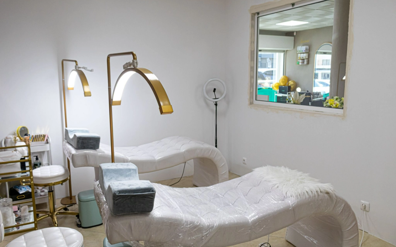 A bright beauty treatment room with two white quilted loungers, tall gold arched lamps and a ring light, with an internal window looking through to the reception.