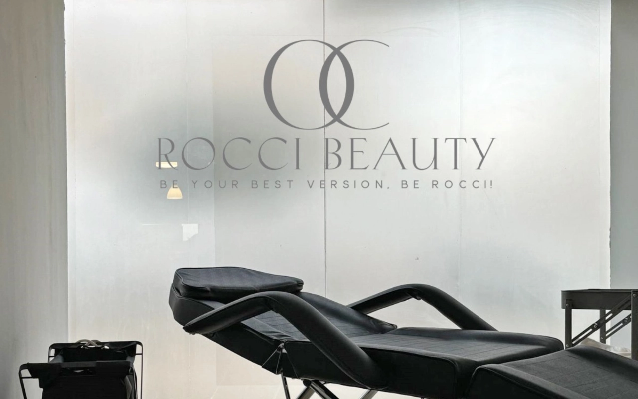 A treatment room with a black reclining couch and a small equipment trolley, set against a frosted-glass wall carrying the Rocci Beauty logo and tagline.