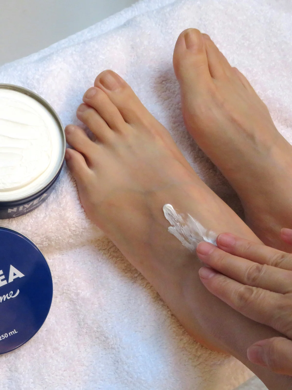 Close-up of the top of a woman’s feet resting on a white sheet with an open pot of moisturiser next to them as she applies moisturiser to her feet