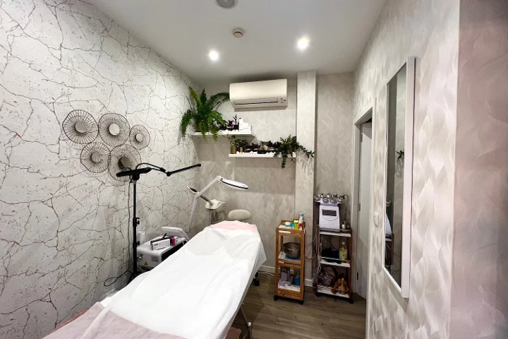 A calm aesthetics treatment room with a covered facial bed, a magnifying lamp and steamer, a trolley of products, textured pale grey walls with decorative metal discs, and trailing plants above the shelving.