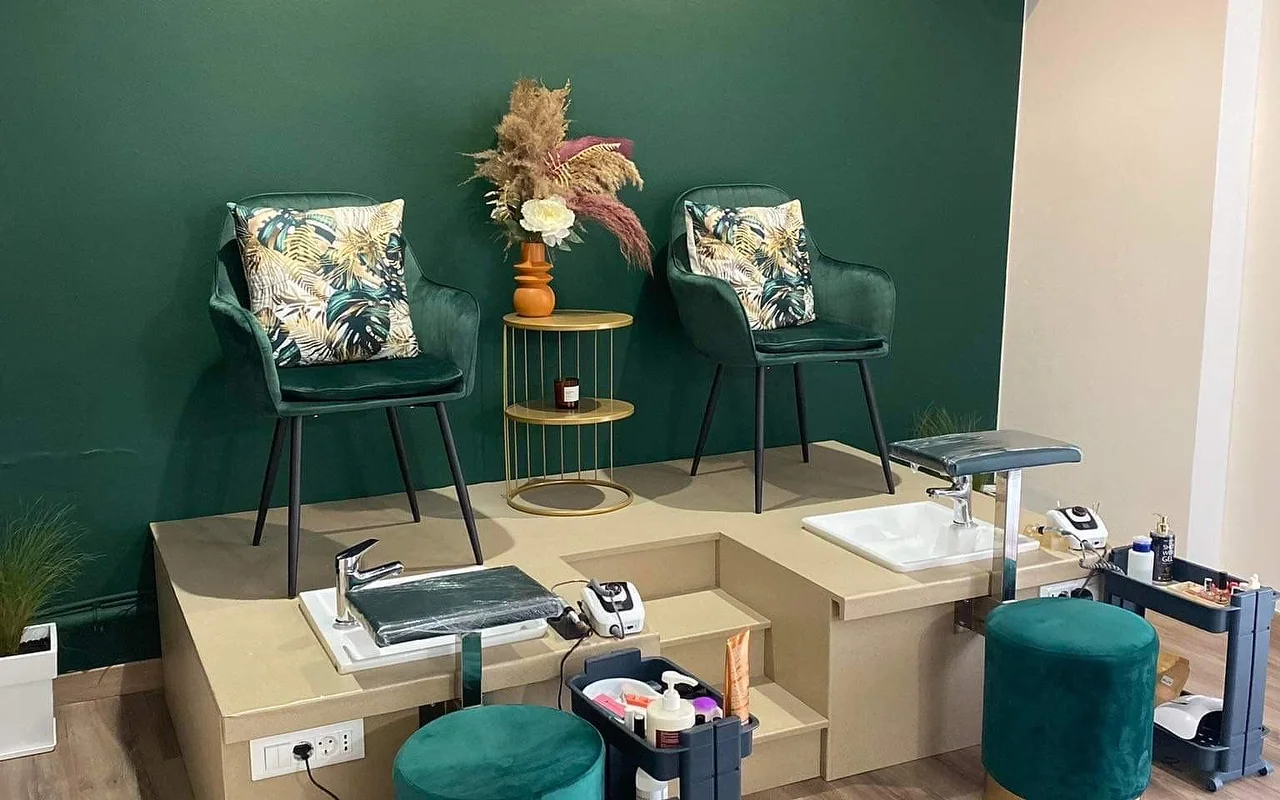 A pedicure corner with a deep green wall, two green velvet tub chairs with tropical-print cushions raised on a beige platform above pedicure basins, a gold side table holding pampas grass in an orange vase, and matching green velvet stools.