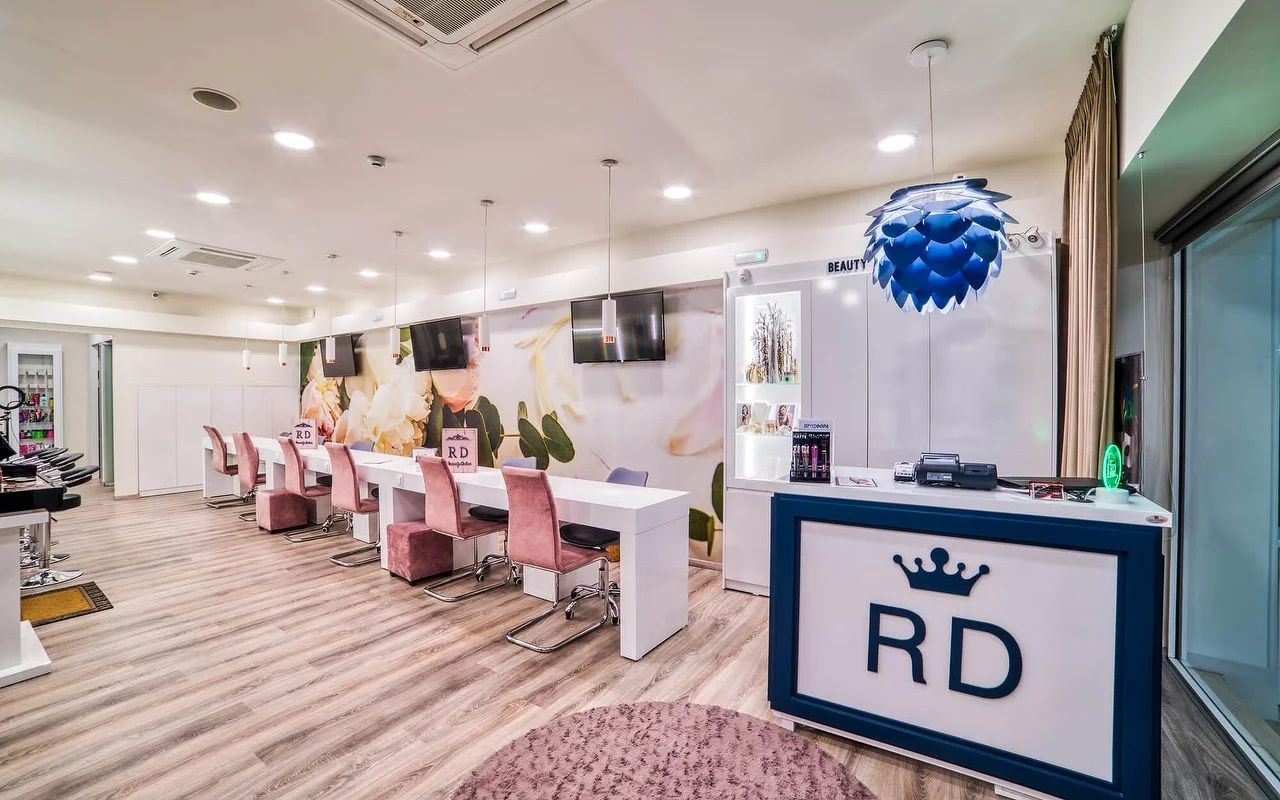 A bright white salon with a long row of manicure tables and dusky-pink chairs, a soft floral wall mural, pale wood flooring, and a reception desk marked with an 'RD' crown logo.