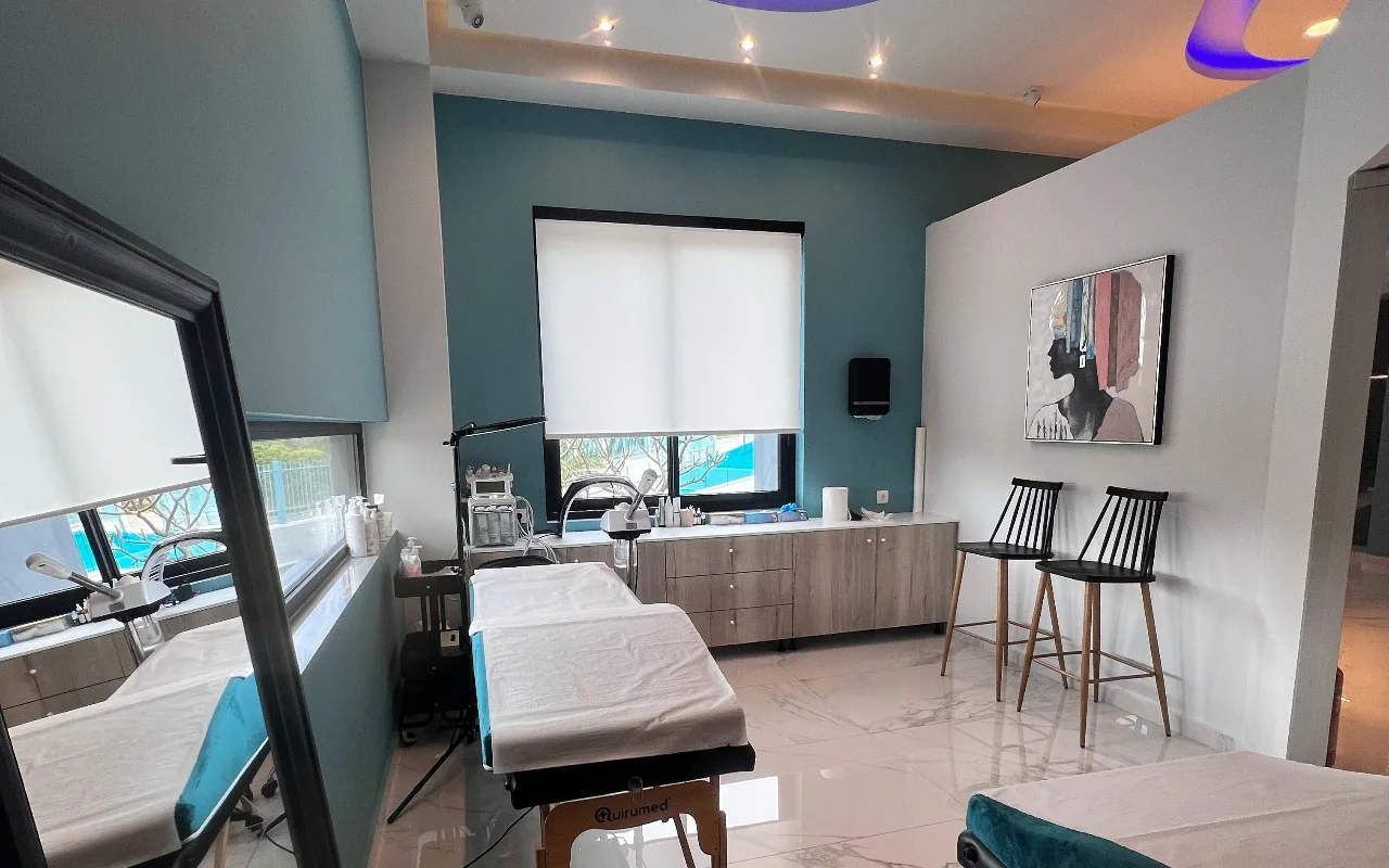 A facial treatment room with teal walls, a white treatment bed, skincare machines, a large mirror, two black slatted stools and a framed portrait, lit by a window with a white roller blind.
