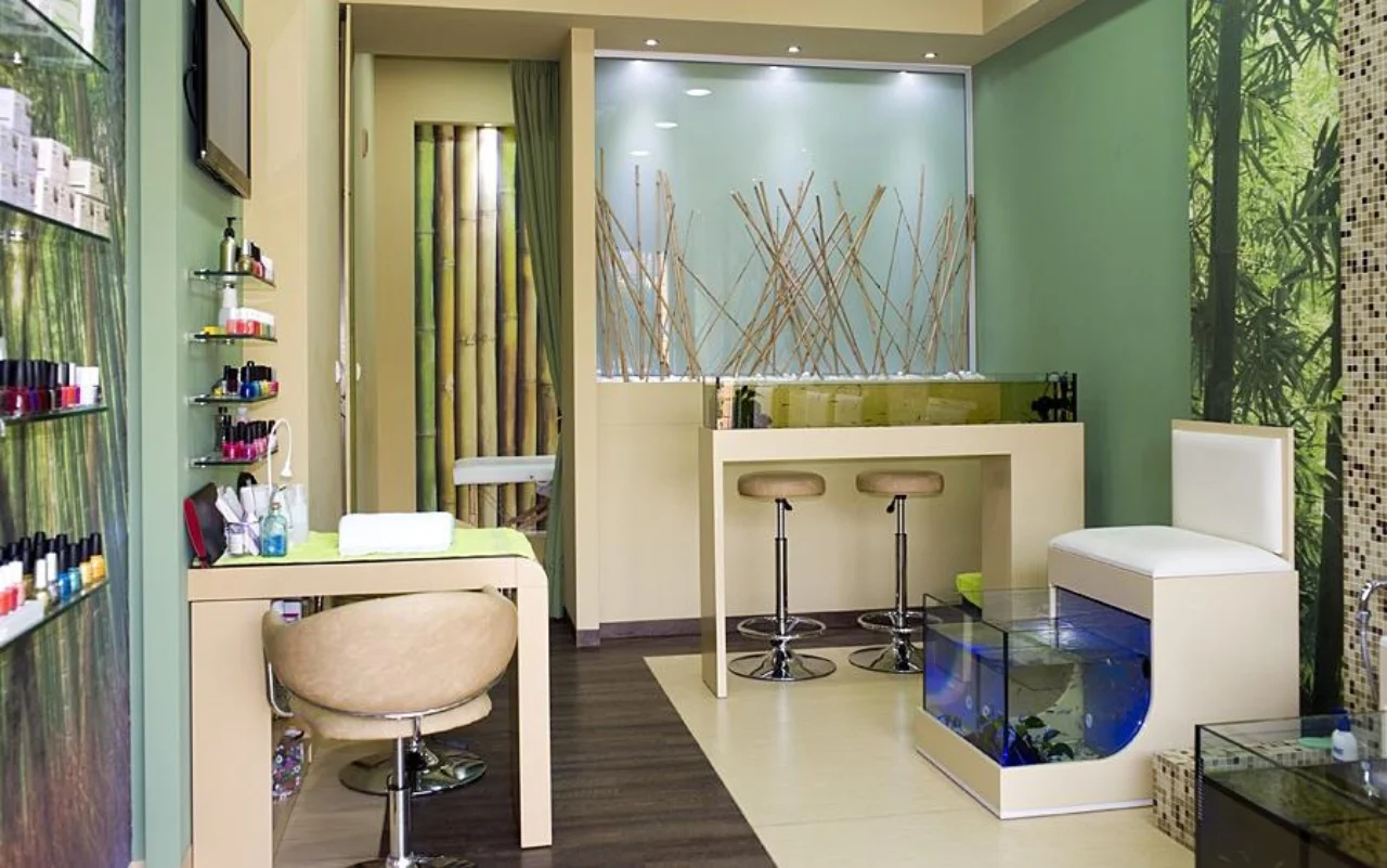 A compact salon with bamboo and green-forest wall decor, wall shelves lined with nail polishes, a cream manicure table and chair, two bar stools, and a blue-lit glass fish tank for the fish-spa pedicure.