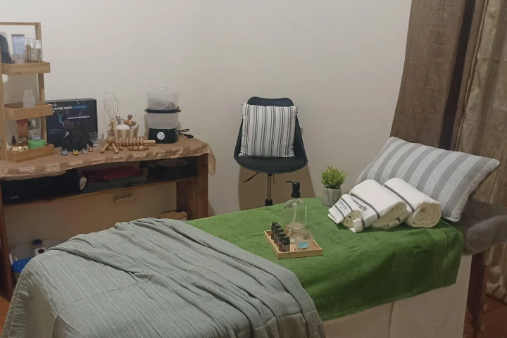 A cosy massage room with a table dressed in a green towel and rolled white towels, a tray of essential oils, wooden massage tools on a rustic side table, and a small stool beside a curtained corner.