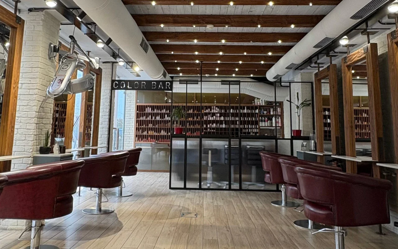 A hair salon with exposed wooden ceiling beams and whitewashed brick, burgundy leather styling chairs, and a glass-walled colour room lined with shelves of product behind a 'Color Bar' sign.
