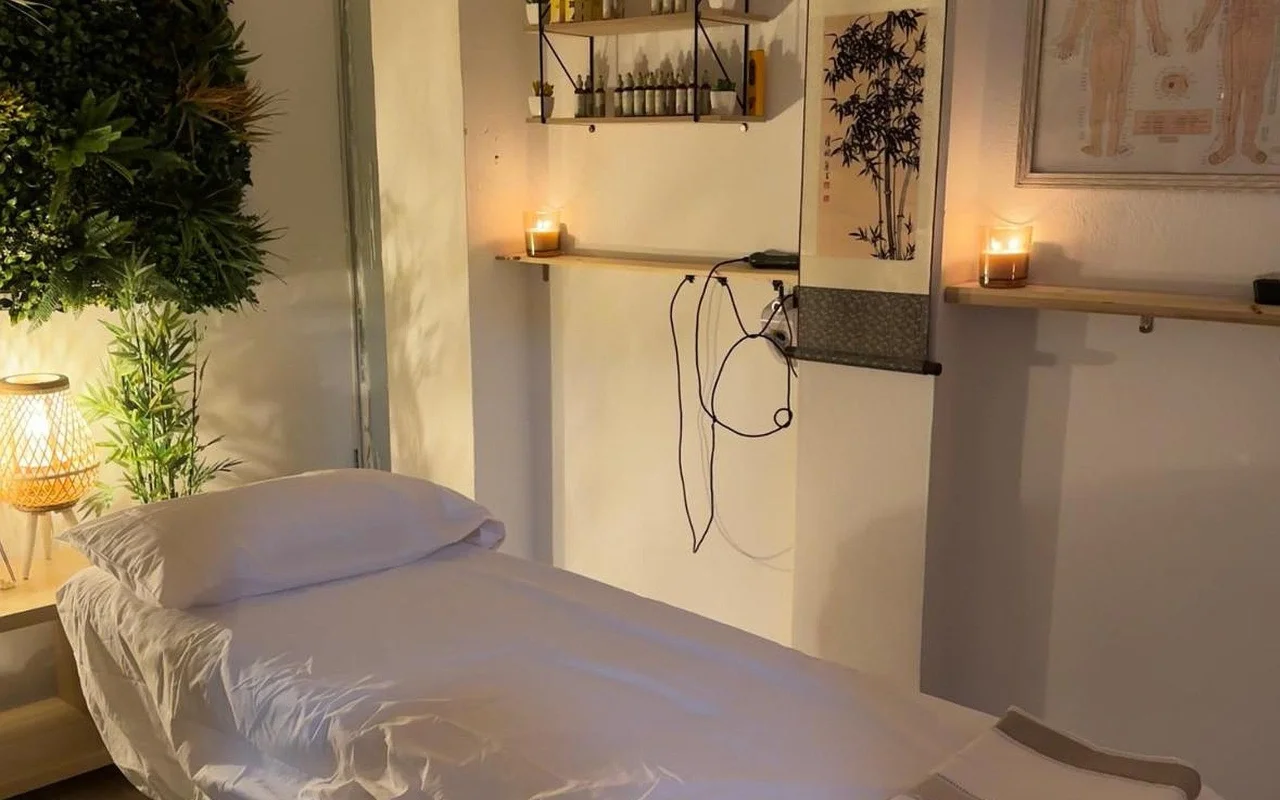 A dimly lit treatment room with a massage bed made up in white linen. Lit candles sit on wooden wall shelves beside a bamboo ink scroll and a framed acupuncture chart, with a leafy plant and a glowing woven lamp in the corner.