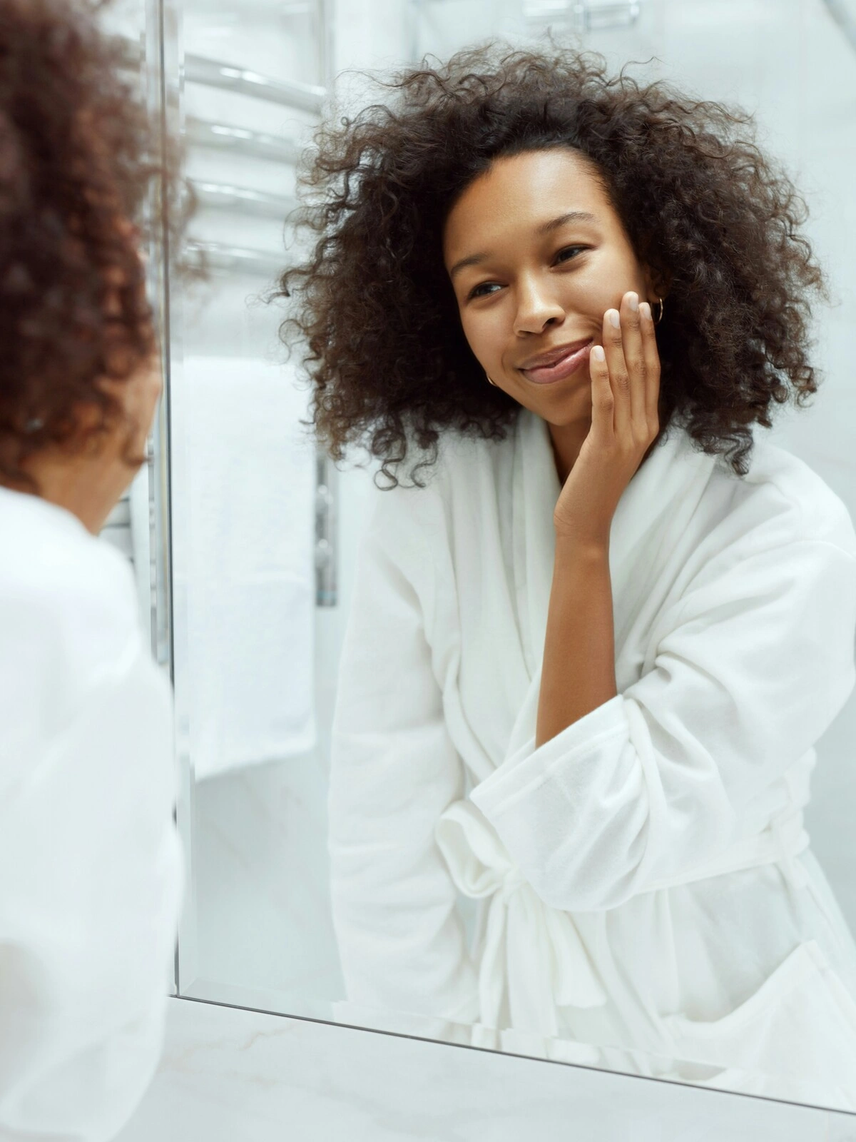 A woman wearing a white bathrobe stands in front of a large mirror in her clean, white bathroom and lightly touches her smooth face