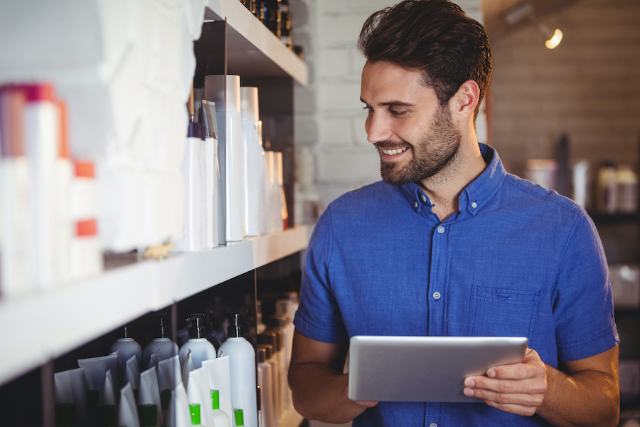Un gérant de salon souriant en chemise bleue consulte une tablette près d'étagères de produits de beauté.