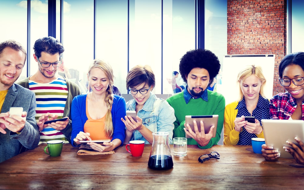 Groupe de personnes diverses autour d'une table utilisant smartphones et tablettes, illustrant la formation en ligne pour coiffeurs et barbiers