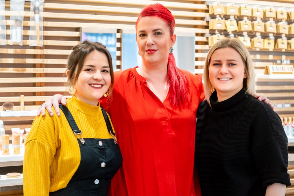 Drie glimlachende salonprofessionals poseren samen met hun armen om elkaar heen voor een houten lattenwand waarop cosmetica staat uitgestald. De vrouw in het midden heeft felrood haar en een rode top.