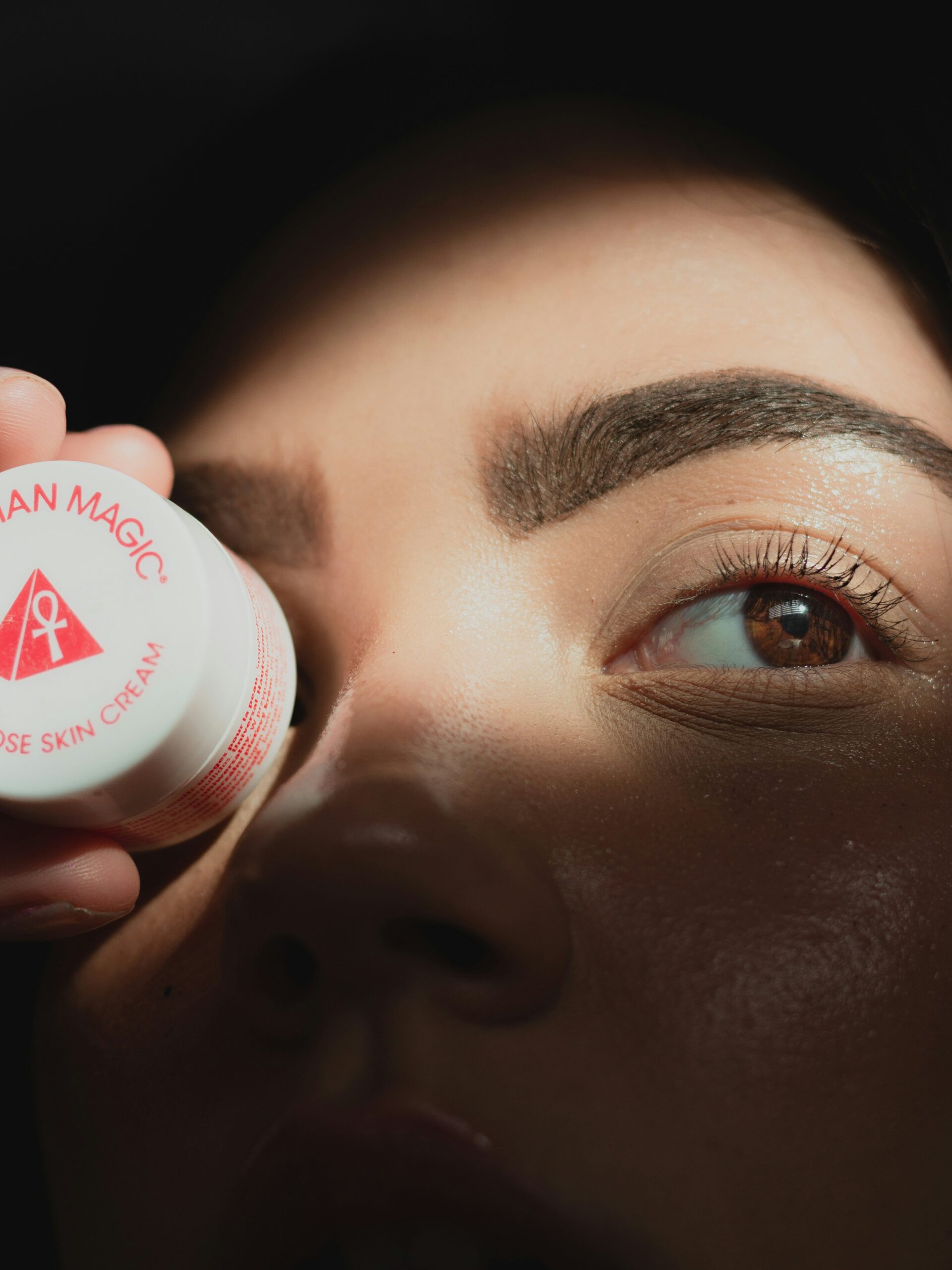 Gros plan sur le visage d’une femme avec un microblading des sourcils, tenant un petit pot de crème rond devant l’un de ses yeux