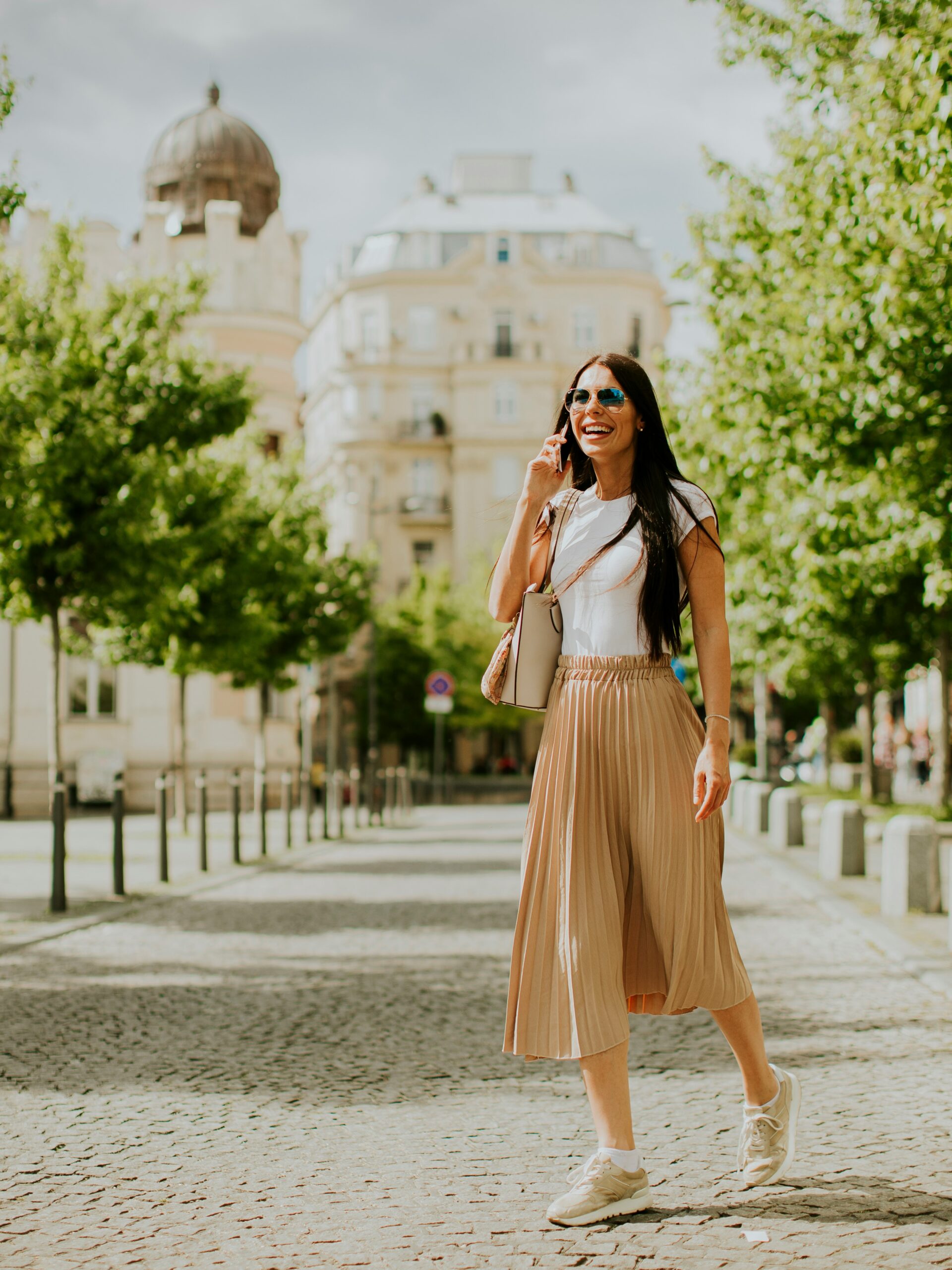 Une femme souriante discute au téléphone en marchant dans une rue pavée, portant un t-shirt blanc ample et une jupe mi-longue fluide