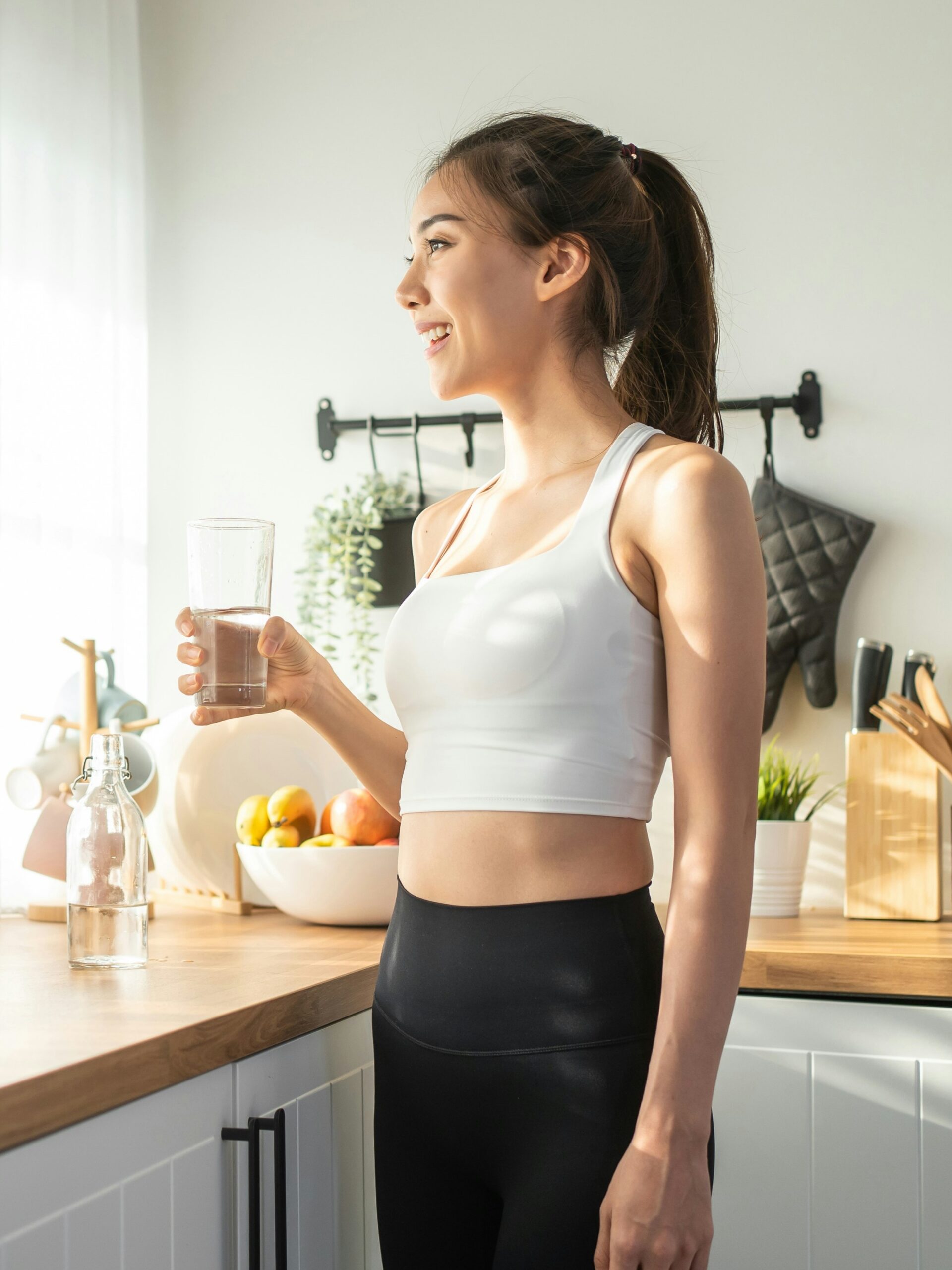 Vue de profil d’une femme buvant un verre d’eau dans sa cuisine devant une fenêtre ensoleillée