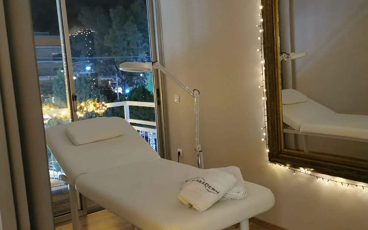 Une salle de soin éclairée en douceur avec un lit blanc, une serviette à l’effigie de CasaDerm, une lampe-loupe, des guirlandes lumineuses encadrant un miroir doré, et une fenêtre avec balcon donnant sur les rues de Glyfáda en soirée.