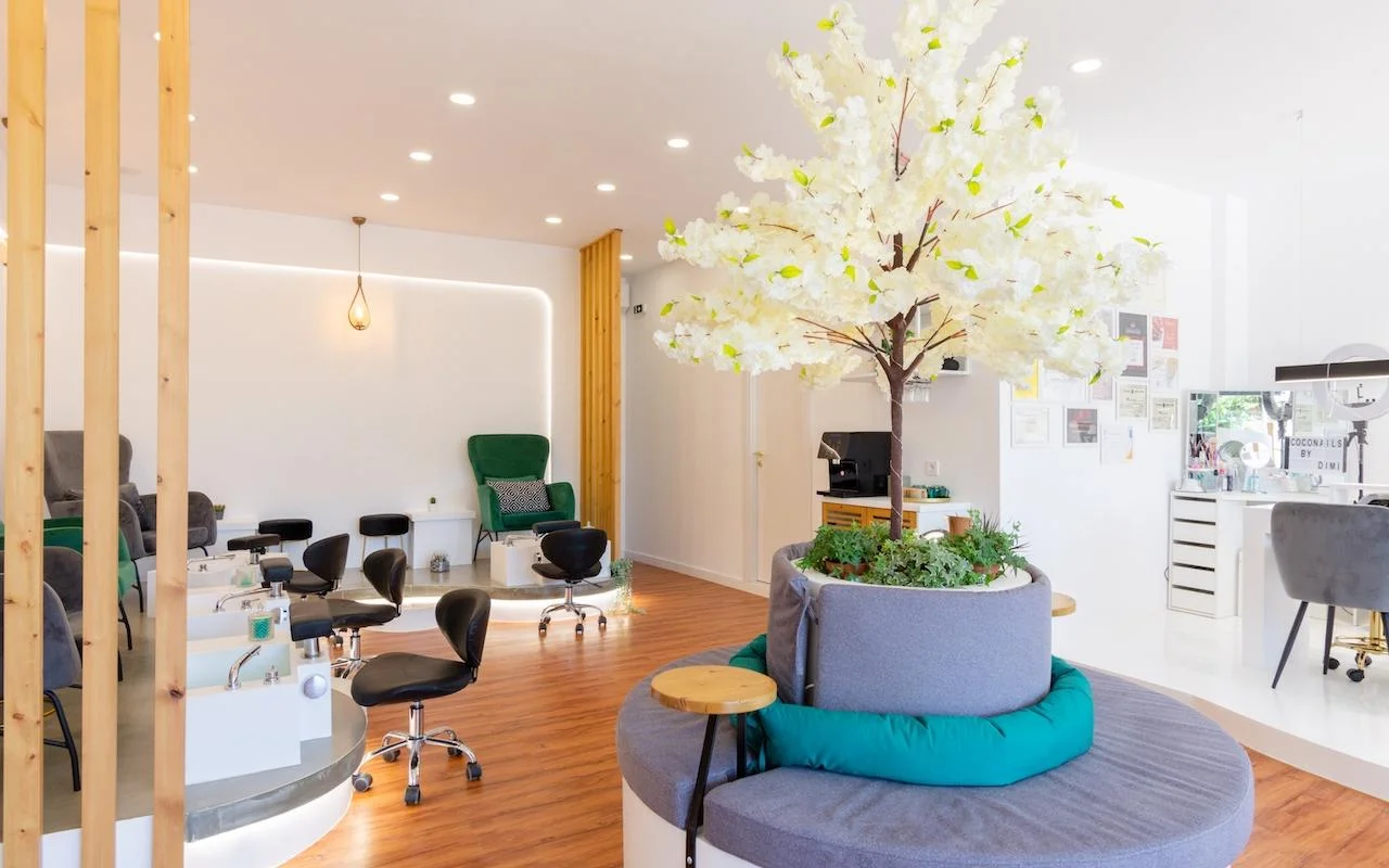 Un salon d’ongles ouvert et inspiré de la nature, avec un grand arbre décoratif à fleurs blanches en pièce maîtresse, des fauteuils en velours vert émeraude aux postes de pédicure, des cloisons en lattes de bois, et un parquet en bois chaleureux.