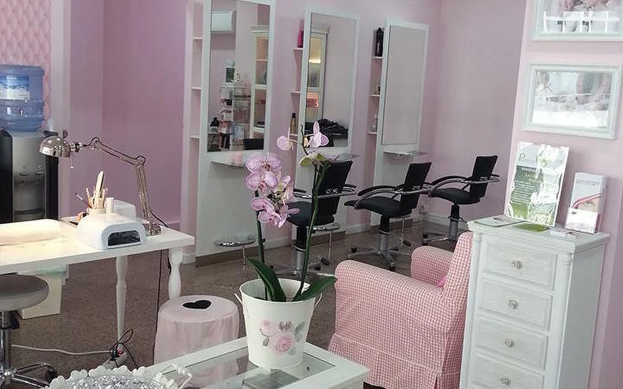 L'intérieur d'un salon de coiffure et d'ongles lumineux aux tons roses. À gauche, un poste de manucure blanc est équipé d'une lampe de bureau et d'un appareil UV. Au centre, une orchidée en pot blanc repose sur une table basse à côté d'un fauteuil vichy rose et blanc. En arrière-plan, plusieurs postes de coiffure avec de grands miroirs et des fauteuils noirs sont disposés contre des murs rose pâle.