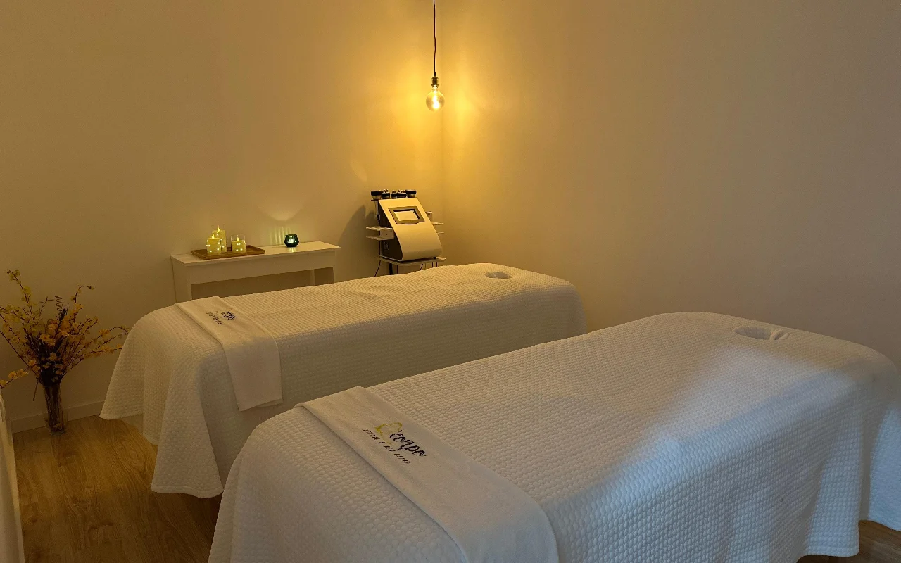 Une cabine de massage ou de soin paisible avec deux tables de soins placées côte à côte, recouvertes de serviettes et de draps blancs impeccables. La pièce est éclairée par une ampoule suspendue et des bougies sur une table d'appoint, créant une atmosphère sereine. Un appareil esthétique spécialisé se dresse en arrière-plan entre les deux lits.
