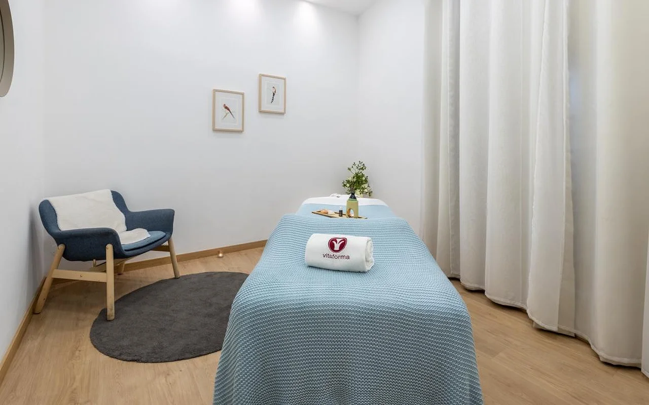 Une cabine de spa épurée et minimaliste aux murs blancs et au parquet clair. Une table de soin est habillée d'un plaid bleu tricoté et d'une serviette blanche roulée portant le logo « Vitaforma ». Dans le coin, un fauteuil bleu élégant repose sur un petit tapis rond gris, et de longs rideaux blancs encadrent la fenêtre.