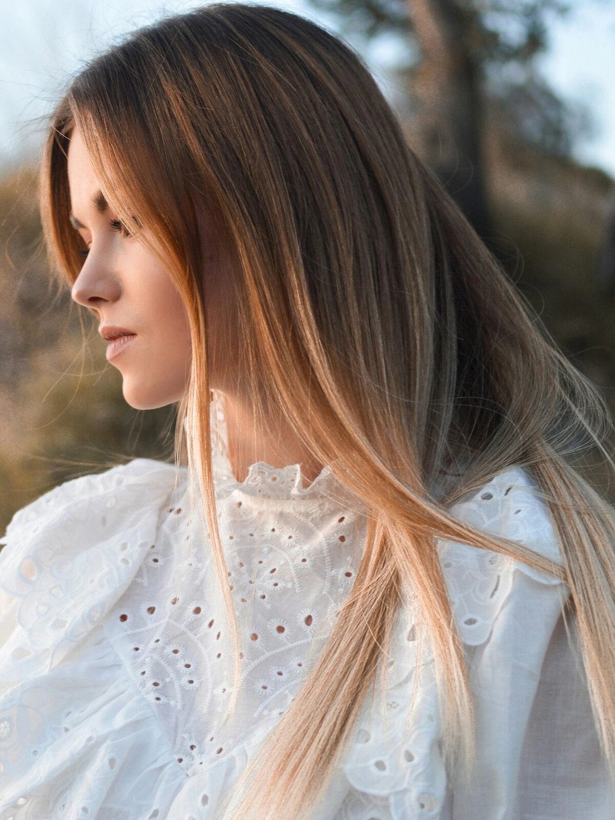 Vue de profil d'une femme en extérieur, dans un cadre naturel, montrant un balayage sur cheveux lisses où le brun se fond dans des nuances blondes