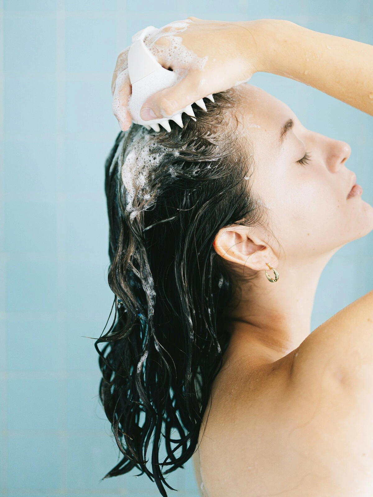 Vue de côté d'une femme aux cheveux mouillés et savonnés massant son cuir chevelu avec une brosse en silicone